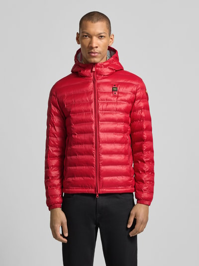 Männermodel trägt Steppjacke mit Zweiwege-Reißverschluss Modell 'Charles' von Blauer Usa in Rot mit Kapuze und Label-Detail, Ganzkörperansicht von vorne. Rückenlänge bei Größe M 65 cm, Schulterbreite 47 cm, Brustweite 55 cm, Ärmellänge 65 cm, Regular Fit.