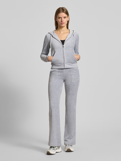 Frauenmodel trägt Sweatjacke mit Label-Stitching Modell 'ROBERTSON' von Juicy Couture, grauer Material-Mix mit Stretch-Anteil, Regular-Fit, Kapuze mit Tunnelzug, Reißverschluss und Känguru-Tasche, Ganzkörperansicht von vorne.
