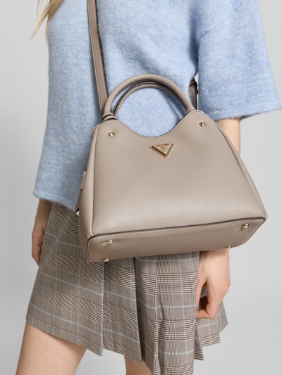 Frauenmodel trägt Handtasche mit Label-Applikation Modell 'ECO MERIDIAN II' von Guess, beige, strukturierte Oberfläche, Hauptfach mit Reißverschluss, Tragehenkel, Schulterriemen, Breite 30 cm, Länge 21 cm, Tiefe 12 cm, Länge Schulterriemen 122 cm, Ganzkörperansicht von vorne.