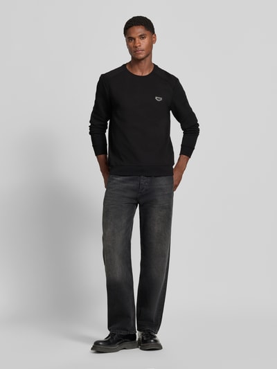 Mannelijk model draagt sweatshirt met labelpatch Model 'NEUTER' van Antony Morato in zwart, getailleerd, ronde hals, elastische boorden, ruglengte maat M 66 cm, mouwlengte maat M 58 cm, versterking op de mouwen. Ganzkörperansicht von vorne.