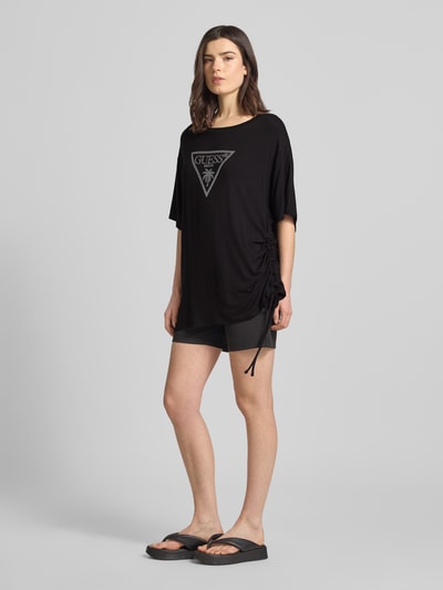 Modelka nosi T-Shirt mit Logo-Print Modell 'COULISSE' von Guess z elastycznego wiskozo-mieszanego materiału, luźny krój, czarny kolor, okrągły dekolt, nadruk z logo, przesunięte ramiona, tunel, długość tyłu w rozmiarze S: 70 cm, pełen widok z przodu.