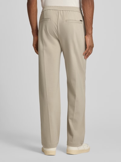Männermodel trägt Slim Fit Hose aus Viskose-Mix Modell 'ALTON' von Guess in Beige mit Bundfalten, Bund und Eingrifftaschen, Ganzkörperansicht Rückansicht.