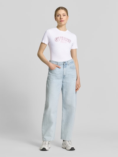 Vrouwenmodel draagt T-Shirt uit viscose-elastaan-mix met logo van Guess Activewear, regular fit, ronde hals, ruglengte maat S 53 cm, wit op lichtblauwe jeans, volledige lichaamsweergave van voren.