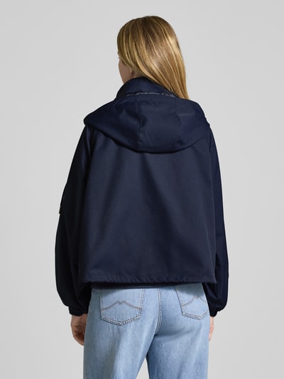 Vrouwenmodel draagt Oversized Jacke mit Kapuze und Zweiwege-Reißverschluss van monari in donkerblauw, glad oppervlak, rugaanzicht, context: dames jack, oversized fit, capuchon, tweewegritssluiting, steekzakken, ruglengte 63 cm, schouderbreedte 33 cm, borstwijdte 59 cm, mouwlengte 53 cm.