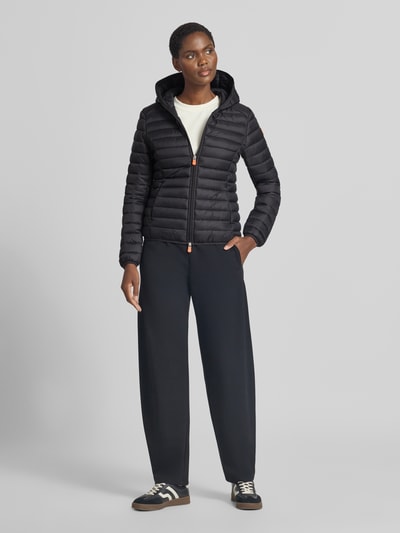 Frauenmodel trägt schwarze Steppjacke mit Zweiwege-Reißverschluss Modell 'DAISY' von Save The Duck, Kapuze, Slim Fit, Ganzkörperansicht von vorne.