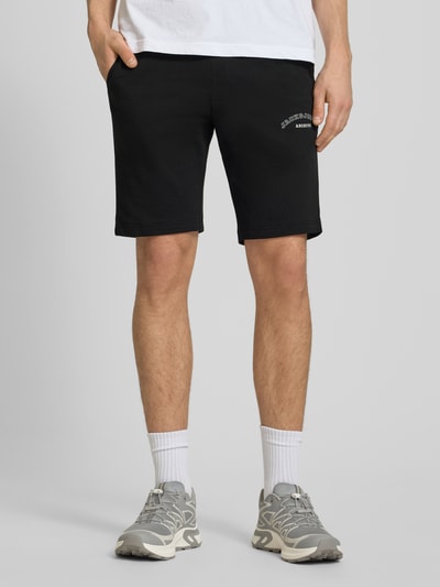 Männermodel trägt schwarze Shorts mit Logo-Print Modell 'GORDON' von Jack & Jones aus Baumwoll-Mix, Relaxed-Fit, Ganzkörperansicht von vorne.