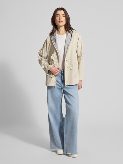 Frauenmodel trägt Regular Fit Regenjacke mit floralen Applikationen Modell 'Rosa' von SAVE THE DUCK in Beige, Kapuze, Reißverschluss, Logo-Patch, weite hellblaue Jeans und weißes Shirt, Ganzkörperansicht von vorne. Rückenlänge bei Größe S: 72 cm, Schulterbreite: 57 cm, Brustweite: 45 cm, Ärmellänge: 50 cm.