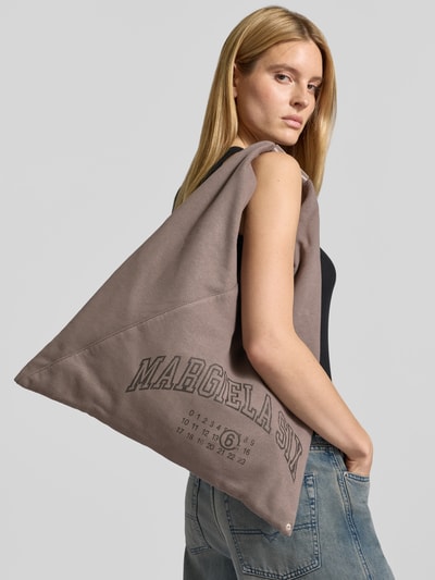 Frauenmodel trägt Handtasche mit Logo-Print von MM6 Maison Margiela in Taupe mit breitem Schulterriemen, Statement-Print, Ganzkörperansicht von vorne.
