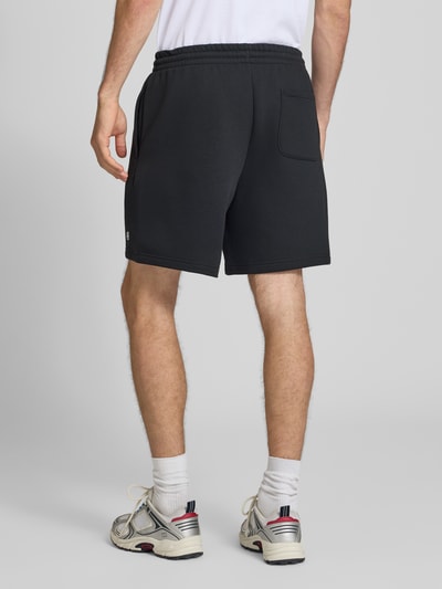 MęskI model nosi Relaxed Fit Shorts mit Logo-Stitching von New Balance, mieszanka bawełny, krój relaxed fit, wyhaftowane logo, elastyczny pas z tunelem, szerokość w rozmiarze M: 42 cm, długość wewnętrzna nogawki w rozmiarze M: 18 cm, widok całej sylwetki z tyłu.