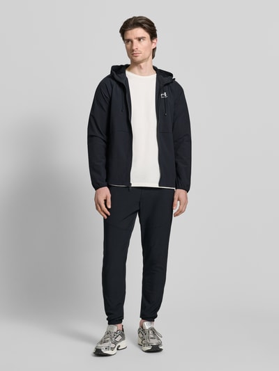 Męski model nosi Kurtkę z kapturem od Under Armour, mieszanka materiałów z dodatkiem streczu, luźny krój, tunel, zamek błyskawiczny, nadruk z logo, wpuszczane kieszenie, długość tyłu w rozmiarze M: 68 cm, długość rękawów w rozmiarze M: 66 cm, czarna, gładka, całkowity widok z przodu.