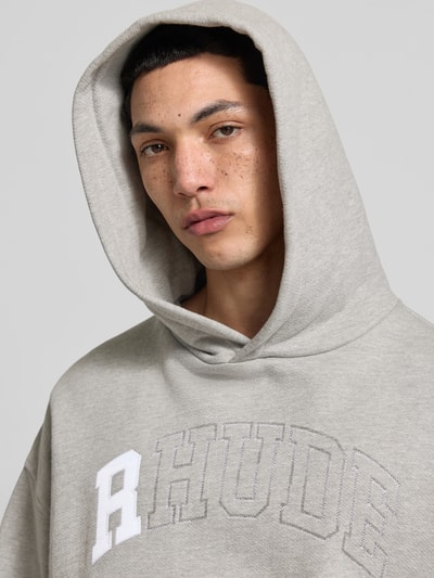 Männermodel trägt Oversized Hoodie mit Logo-Stitching von Rhude aus Baumwoll-Mix in hellem Grau, Nahaufnahme Oberkörper.