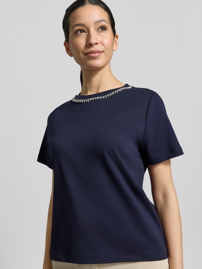 Kobiece model prezentuje t-shirt z bawełny w kolorze granatowym, perły i obszycie ozdobnymi kamieniami, krój regular fit, okrągły dekolt, długość tyłu w rozmiarze S: 53 cm, T-Shirt mit Perlen und Ziersteinbesatz od Christian Berg Woman, półzbliżenie na górną część ciała.