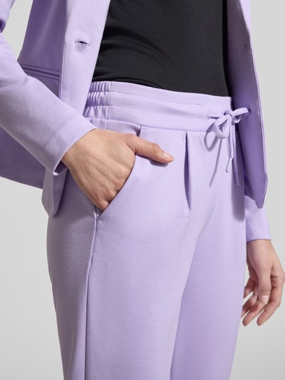 Kobieta nosi Slim Fit Stoffhose mit Viskose-Anteil Modell 'Kate' od ICHI, kolor jasnofioletowy, elastyczny materiał, krój slim fit, szerokość w rozmiarze S: 38 cm, długość nogawki od wewnątrz: 65 cm, kieszenie, wiązany pas, detal spodni, zbliżenie na biodra