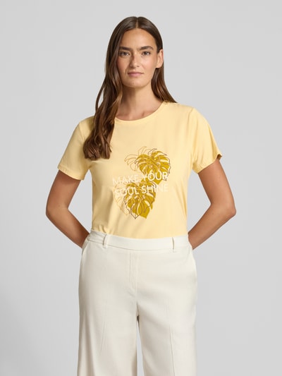 Kobiece model nosi damski t-shirt z bawełny, krój regular fit, okrągły dekolt, nadruk z napisem i motywem, model 'RINA' od Soyaconcept, długość tyłu 60 cm w rozmiarze S, jasny żółty, przód z motywem; zdjęcie całej sylwetki z przodu.