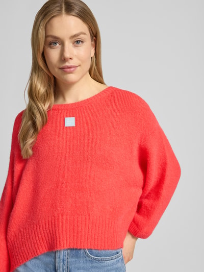 Frauenmodel trägt Relaxed Fit Pullover mit Alpaka-Anteil Modell 'East' von American Vintage in Rot, Strick-Optik, Statement-Detail, U-Boot-Ausschnitt, elastische Abschlüsse, Elastischer-Material-Mix, Rückenlänge 42 cm, Ärmellänge 42 cm, Ganzkörperansicht von vorne.