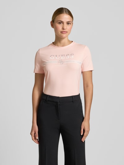 Frauenmodel trägt T-Shirt mit Strasssteinbesatz Modell 'SIENA' von Guess, rosa, Logo-Print, Baumwoll-Mix, Ganzkörperansicht von vorne.