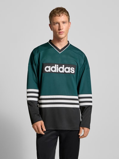 Männermodel trägt Longsleeve mit Label-Detail von adidas Originals in Grün und Schwarz, Lochstickerei, V-Ausschnitt, Lockerer Schnitt, Ganzkörperansicht von vorne. Rückenlänge bei Größe M: 76 cm.