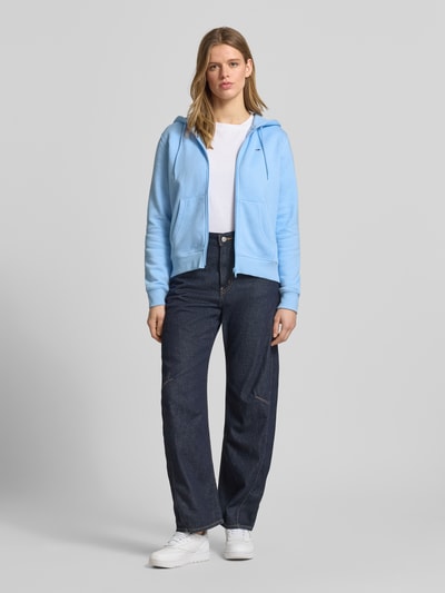 Frauenmodel trägt Regular Fit Sweatjacke aus Baumwoll-Mix von Tommy Jeans in Hellblau, unifarben, Kängurutasche, Ganzkörperansicht von vorne.
