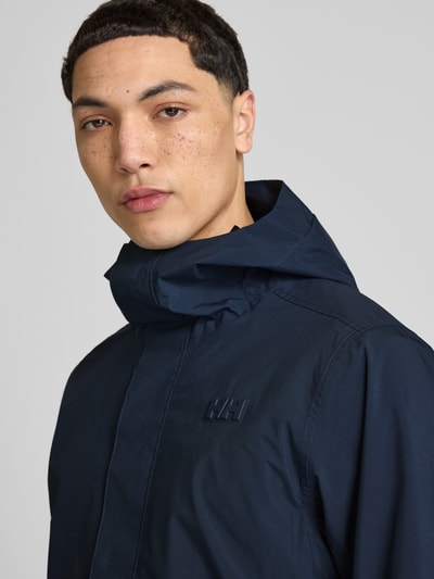 Męski model nosi Regular Fit Regenmantel mit Logo-Detail Modell 'Brussel' von Helly Hansen, ciemnogranatowy kolor, detal z logo, kaptur, wpuszczane kieszenie, stójka, długość tyłu w rozmiarze M 90 cm, szerokość ramion w rozmiarze M 42 cm, szerokość klatki piersiowej w rozmiarze M 51 cm, długość rękawów w rozmiarze M 68 cm, zbliżenie na górną część torsu