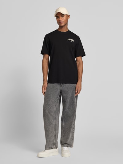 Męski model nosi T-Shirt z label-printem model 'Brady whop' od Les Deux, bawełna, krój regular fit, czarny, widok całej sylwetki z przodu.