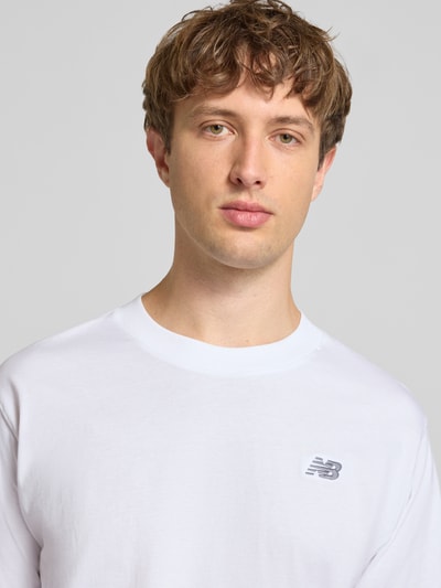 Męskimodel nosi T-Shirt mit Logo und Rundhalsausschnitt von New Balance z czystej bawełny, krój regular fit, logo, okrągły dekolt, długość tyłu w rozmiarze M: 74 cm, kolor biały, faktura gładka, zbliżenie na górną część ciała, przód.