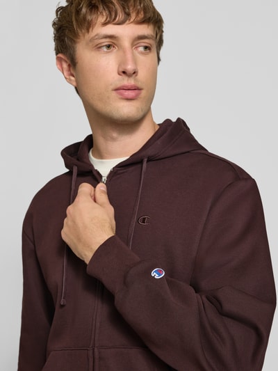 Männermodel trägt Sweatjacke mit Label-Stitching und Känguru-Tasche von Champion aus Baumwoll-Mix in Dunkelbraun, Nahaufnahme Oberkörper.