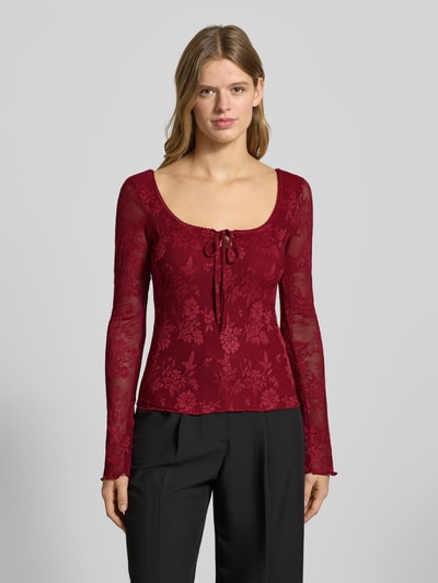 Modelka nosi semitransparentne longsleeve z wiązaniem od Gina Tricot, materiał z domieszką stretchu, slim fit, karo dekolt, przód na całej długości, kolor bordowy z roślinnym wzorem, długość tyłu S: 29 cm, zdjęcie całej sylwetki z przodu.