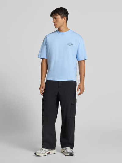 Männermodel trägt Regular Fit T-Shirt aus reiner Baumwolle mit rückseitigem Print von REVIEW, hellblau mit Logo-Detail, Ganzkörperansicht von vorne. Rückenlänge bei Größe M: 67 cm.