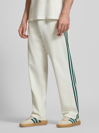 Männermodel trägt Sweatpants mit Reißverschlusstaschen und Logo-Stitching von adidas Originals in Weiß mit grünen Streifen, lockerer Schnitt, Nahaufnahme Unterkörper.