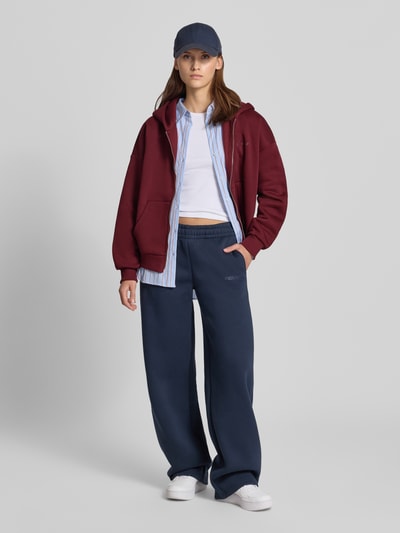 Kobiece modelka nosi Relaxed Fit Sweatjacke aus Baumwoll-Mix mit Label-Detail od REVIEW, bordowy kolor, z elastycznymi ściągaczami, osobna kieszeń kangurka, zamek błyskawiczny, luźny krój, tekstura miękka, widoczne detale marki, widok całej sylwetki z przodu.
