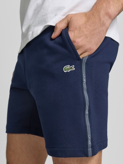 Männermodel trägt Regular Fit Sweatshorts aus reiner Baumwolle von Lacoste in Dunkelblau mit Logostreifen, Nahaufnahme Oberkörper.