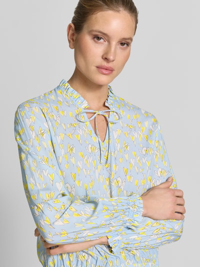 Frauenmodel trägt Regular Fit Bluse aus reiner Viskose mit elastischen Abschlüssen von Emily Van den Bergh, hellblau mit gelbem und weißem Allover-Motiv, Stehkragen mit Rüschenbesatz, Schlüsselloch-Ausschnitt, feine Textur, Langarm, Ganzkörperansicht von vorne.