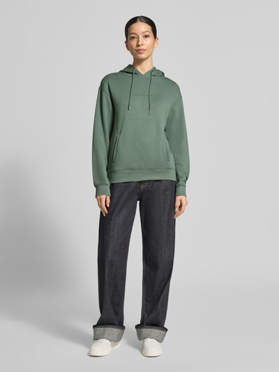 Modelka kobieca nosi Hoodie z tunelem model 'IMA' od MSCH Copenhagen, zielony melanż, prosty krój, kieszenie, całościowy widok z przodu.