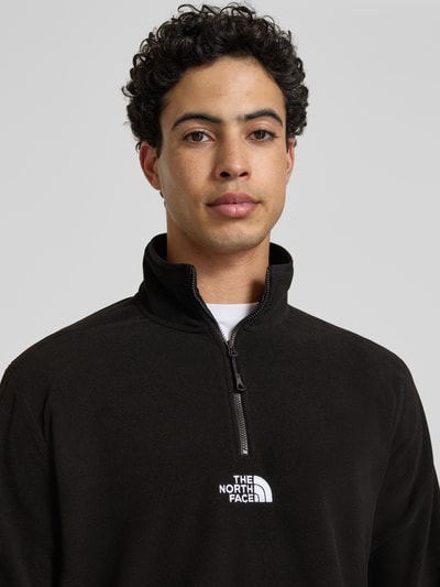 Männermodel trägt schwarzen Fleecepullover mit Reißverschluss Modell 'GLACIER' von The North Face, Label-Stitching, Nahaufnahme Oberkörper.