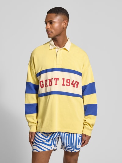 Męski model nosi Relaxed Fit Longsleeve mit Logo-Print od Gant, bawełna, krój relaxed fit, logo-print, kołnierzyk polo, elastyczne mankiety, długość tyłu w rozmiarze M 79 cm, długość rękawa w rozmiarze M 64 cm, żółty z niebieskimi paskami i czerwonym logo, widok całej sylwetki z przodu.