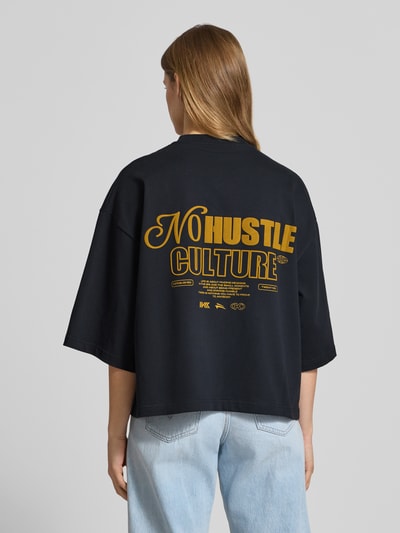 Frauenmodel trägt Cropped T-Shirt mit rückseitigem Statement-Print von Karo Kauer in Schwarz mit gelbem Statement-Print, Reine Baumwolle, Loose-Fit, Logo-Print, Ganzkörperansicht Rückansicht.