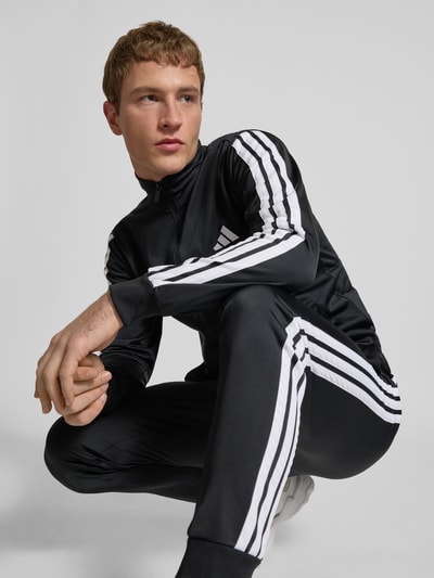 Männermodel trägt Trainingsanzug mit Stretch-Anteil von adidas Sportswear, elastyczna mieszanka materiałów, prosty krój, stójka, zamek błyskawiczny, elastyczne wykończenia, elastyczny pas, wpuszczane kieszenie, detale z logo, szerokość w rozmiarze M: 39 cm, długość tyłu w rozmiarze M: 70 cm, szerokość ramion w rozmiarze M: 44 cm, długość rękawów w rozmiarze M: 65 cm, długość wewnętrzna nogawki w rozmiarze M: 73 cm, czarny z białymi paskami, gładka tekstura. Perspektywa: całkowita sylwetka z przodu.