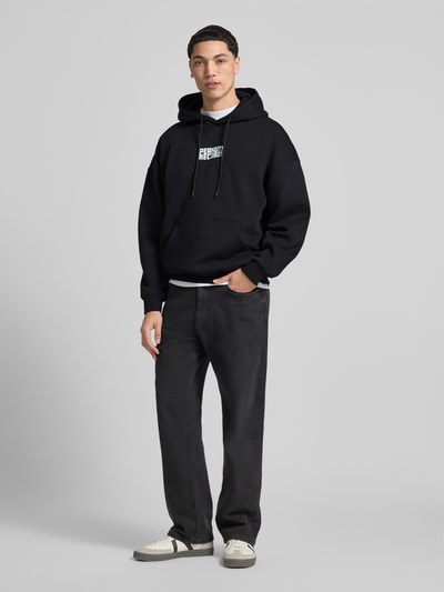 Męski model nosi Oversized Hoodie aus Baumwoll-Mix Modell 'PEANUTS BACK' od Only & Sons, czarny, nadruk peanuts®, kieszeń kangurka, dzianina dresowa, krój oversized, długi rękaw, przód, widok całej sylwetki.