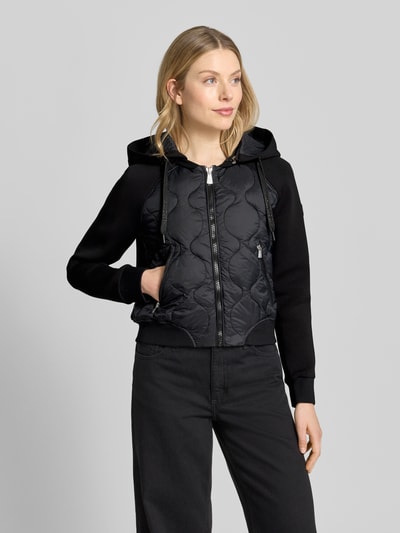 Frauenmodel trägt Steppjacke mit Reißverschlusstaschen Modell 'MALAYA' von khujo, schwarz, Regular-Fit, Kapuze, Ganzkörperansicht von vorne, Rückenlänge bei Größe S 50 cm, Schulterbreite 47 cm, Brustweite 49 cm, Ärmellänge 71 cm.