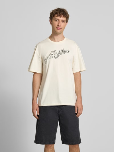Męski model nosi Oversized T-Shirt mit Logo-Stitching von KARL KANI, czysta bawełna, krój oversized fit, okrągły dekolt, długość tyłu w rozmiarze M: 73 cm, jasnokremowy z czarnym logo, widok całej sylwetki od przodu.