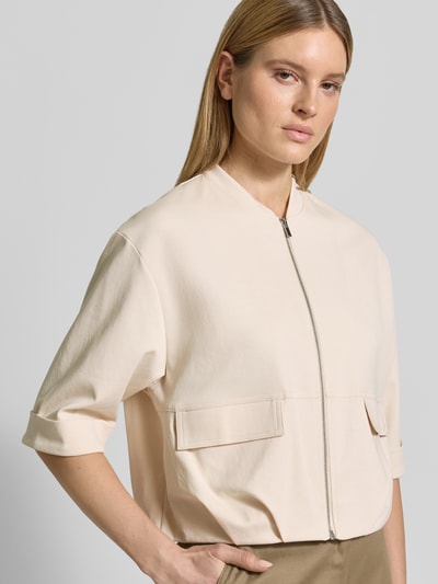Frauenmodel trägt Blouson mit Reißverschluss und Pattentaschen Modell 'Goros' von OPUS aus Baumwoll-Mix mit Stretch-Anteil, Relaxed-Fit, Stehkragen, 3/4-Arm, Blusen-Optik in Creme mit glatter Textur, Nahaufnahme Oberkörper.
