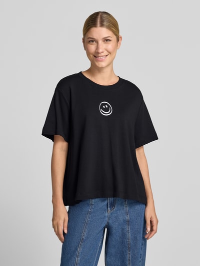 Kobiece model prezentuje czarny Damski t-shirt Boxy Fit T-Shirt aus reiner Baumwolle model 'SARA' od VERO MODA z białą grafiką, całkowity widok z przodu.