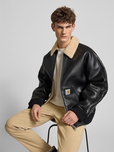 Männermodel trägt schwarze Bomberjacke mit Zweiwege-Reißverschluss Modell 'SHEPTON' von Carhartt, Teddykragen, Nahaufnahme Oberkörper. Relaxed Fit, Eingrifftaschen, Label-Detail, Materialstruktur sichtbar.