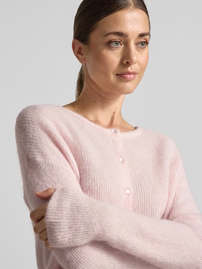Frauenmodel trägt Strickjacke aus Alpaka-Mix von MOS MOSH in zartem Rosa, gerippter Rundhalsausschnitt, Nahaufnahme Oberkörper, lockere Passform, weiche Textur, KI-modifiziertes Bild.