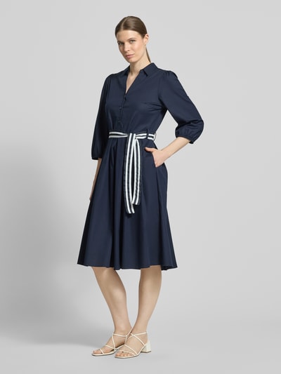 Frauenmodel trägt Hemdblusenkleid mit V-Ausschnitt und Bindegürtel von More & More aus dunkelblauem Baumwoll-Elasthan-Mix, A-Linie, Midikleid, mit Knopfleiste, Rückenlänge 109 cm, Brustweite 47 cm, Ärmellänge 49 cm, Ganzkörperansicht von vorne.
