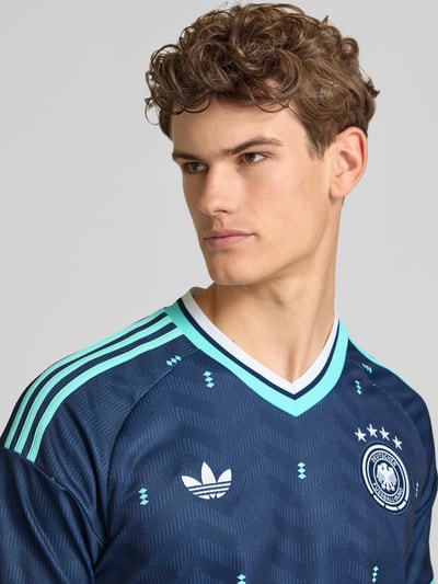Männermodel trägt DFB Auswärtstrikot Deutschland 2026 von adidas Sportswear, Mehrfarbig, Slim-Fit, Logo-Applikation, V-Ausschnitt, Ärmelform: Kurzarm, Nahaufnahme Oberkörper.