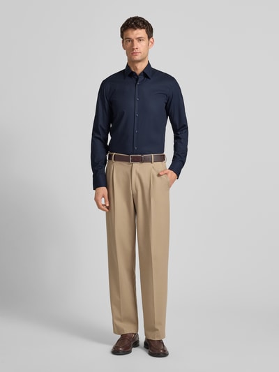 Męski model nosi Slim Fit Businesshemd aus Popeline mit Stretch-Anteil od BOSS, mieszanka bawełny i elastanu, krój slim fit, kołnierzyk typu kent, na całej długości listwa guzikowa, sportowe mankiety, zaokrąglony dół, łatwa pielęgnacja, długość tyłu w rozmiarze 40: 76 cm, szerokość ramion w rozmiarze 40: 47 cm, długość rękawów w rozmiarze 40: 67 cm, granatowa góra, beżowe spodnie, brązowy pasek, brązowe buty, gładka struktura, pełny widok z przodu.