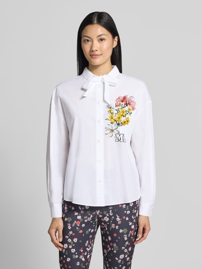 Frauenmodel trägt Relaxed Fit Hemdbluse aus reiner Baumwolle von Marc Cain, weiß mit buntem Blumen-Print, Langarm, Logo-Print, Hemdblusenkragen, Brusttasche; man erkennt Stehkragen mit Tunnelzug und lockeren Schnitt; Ganzkörperansicht von vorne.