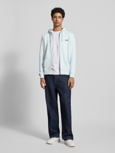 Męski model nosi Regular Fit Sweatjacke mit Logo-Stitching od Superdry z mieszanki bawełny, jasnoniebieska, przód, krój regular fit, kaptur z tunelem, zamek błyskawiczny, kieszeń kangurka, elastyczne wykończenia, długość tyłu 69 cm, długość rękawów 65 cm, widok całej sylwetki z przodu.