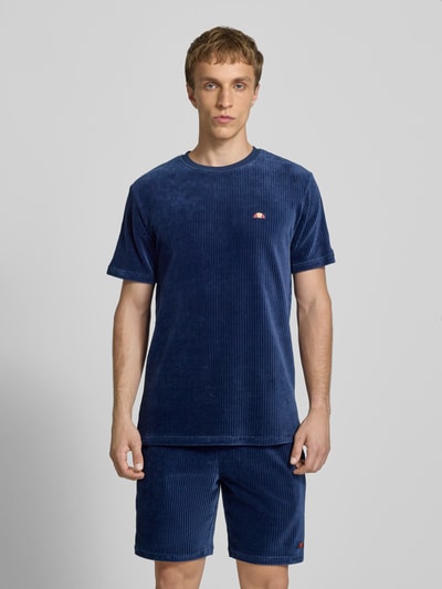 Męski model nosi T-Shirt mit Logo Modell 'MADORI' od Ellesse, mieszanka bawełny z dodatkiem streczu, krój regular fit, okrągły dekolt, logo, długość tyłu w rozmiarze M: 66 cm, kolor granatowy, prążkowana tekstura, widok całej sylwetki z przodu.